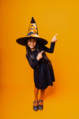 Little cheerful Caucasian girl in a Halloween witch costume points her finger to the side.