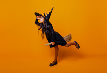 Little girl dressed as a Halloween witch in a black dress and hat flies on a broomstick on a yellow...