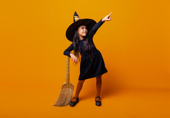 Little girl dressed as a Halloween witch in a black dress and hat flies on a broomstick on a yellow...