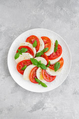 Classic caprese salad with mozzarella cheese, tomatoes and basil on a white plate on a gray concrete background. Top view, copy space.