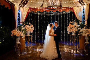 newlyweds at evening wedding ceremony in park among light bulbs and garlands.