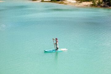 A girl in a dress floats on a glanders board on a pond with bright turquoise water. Warm summer day for travel. 