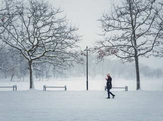 Lone young woman walking in the park drinking a hot coffee in a cold winter day. Seasonal blizzard weather on the street. Snowfall morning in the square. Gils wandering under snowy trees