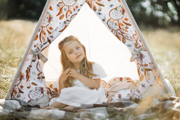 Adorable little blond girl sitting in boho teepee tent. Cute girl child, wearing white dress and feather hair accessories in natice american style, posing to camera in wigwam outdoors in summer field © sofiko14