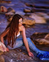 Beautiful fashionable mermaid with red hair holding shell sitting on rock near sea