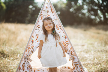 Summer wild field and forest, happy childhood concept. Pretty cute little blond girl dressed in indian american style, standing in front of teepee wigwam with sleep catchers, outdoors at sunset © Sofiia Shunkina