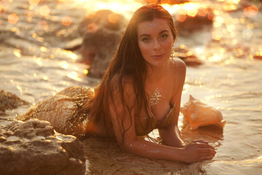 Beautiful fashionable mermaid with red hair holding shell sitting on rock near sea