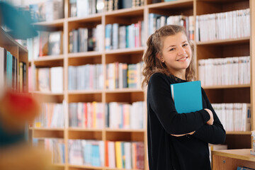 Happy tenage girl or smiling student holding a blue book wih copy space among many books in library - People, knowledge, education and school concept