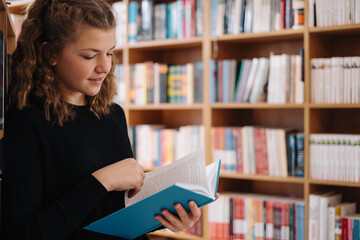 Teen girl among a pile of books. A young girl reads a book with shelves in the background. She is surrounded by stacks of books. Book day.