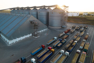 Grain terminals of modern sea commercial port. Silos for storing grain in rays of setting sun. Many trucks are waiting in line for unloading in port harbor, top view from quadcopter. Logistics, trade - obrazy, fototapety, plakaty