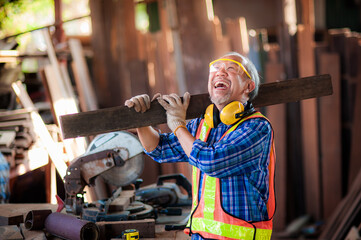 Laughing and happy Asian elderly carpenter craftsman in carpentry shop wearing work clothes with work safety equipment such as gloves, noise protection equipment and carried a plank get ready to work.