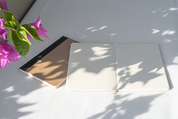 Modern summer stationery still life. Blank diary, notepad mockup with bougainvillea branch in the foreground in sunlight. Long shadows of the sunset on the open pages and on the white tablecloth. 