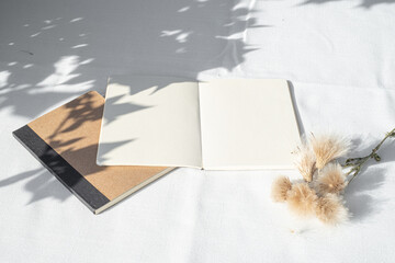 Modern summer stationery still life. Blank diary, notepad mockup with floral element in sunlight. Long shadows of the sunset on the open pages and on the white tablecloth. Top view