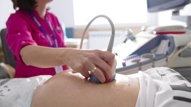 Female doctor examining belly of pregnant woman with ultrasound. Gynecologist moving transducer on abdomen of a future mother in clinic during ultrasonography process.