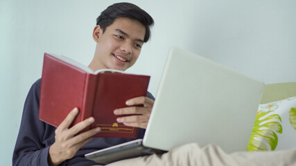 young asian man holding book happily while looking at laptop screen at home