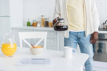 cropped view of african american man holding coffee pot while looking at breakfast on table in kitchen