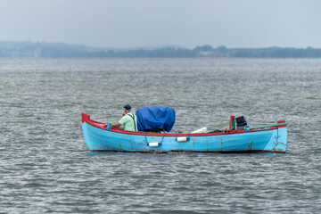 Fisherman in green t-shirt smoking a cigarette sits in front of his small blue boat, sailing into Baltic sea near Jastarnia, Poland, on a cloudy summer day. Land in the haze on distant horizon.