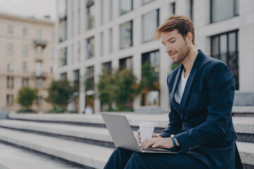 Businessman in formal suit reads news via laptop computer works online drinks takeaway coffee
