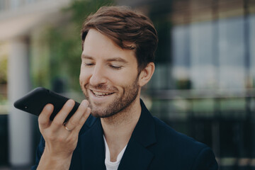 Handsome smiling neat bearded office worker talking on speakerphone standing outside