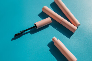 pink clean makeup brush for mascara lies next to an open tube, closed tubes of cosmetics, lip gloss, liquid lipstick, pink eyeliner on a blue background with shadows. Copy space