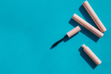 pink clean makeup brush for mascara lies next to an open tube, closed tubes of cosmetics, lip gloss, liquid lipstick, pink eyeliner on a blue background with shadows. Copy space