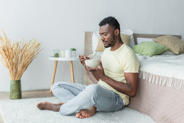 bearded african american man sitting with crossed legs near bed while having breakfast