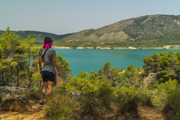 Fototapeta premium mujer haciendo ruta de senderismo, caminar en las montañas, mujer de pelo rosa subida a una roca mirando el lago, mujer joven mirando el embalse desde la montaña