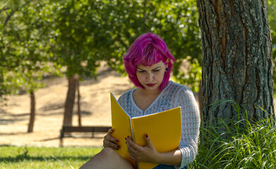 mujer de pelo rosa estudiando sentada junto a un árbol en el parque, mujer de pelo corto con un cuaderno amarillo en la mano, mujer de pelo rosa leyendo junto a un árbol - obrazy, fototapety, plakaty
