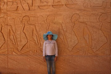 A young girl with a hat standing in front of an ancient wall in Abou simbel temple featuring prisoners in Aswan in Egypt - obrazy, fototapety, plakaty