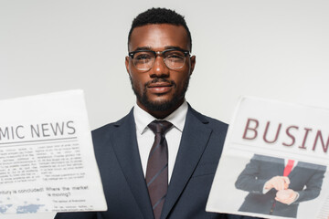 african american man in eyeglasses holding newspapers isolated on grey