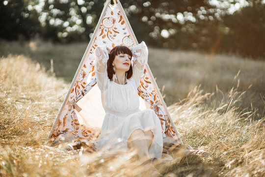 Harmony with nature, meditation, calm and ethnic people. Young relaxed ethnic native american woman sitting in front of teepee wigwam, with arms behind head and eyes closed