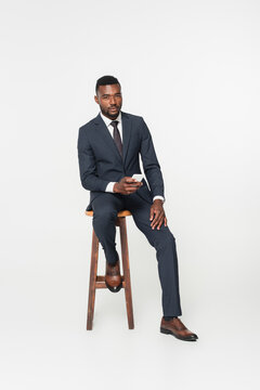 full length of african american businessman in suit sitting on wooden stool and using smartphone isolated on grey