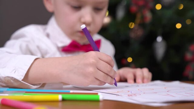 Boy learns how to draw and talks about his drawings, or paints greeting card for parents for Christmas, writes letter with wishes to Santa, hand with felt-tip pen in foreground, blurred background.