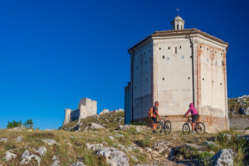 Man and woman riding mtb to castle ruins on mountain top at Rocca Calascio, italian travel destination, Gran Sasso National Park, Abruzzo, Italy. Clear blue sky