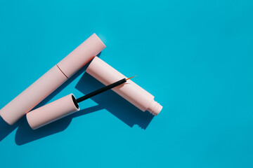 pink makeup brush for eyeliner lies on an open tube, closed tubes of cosmetics, mascara, lip gloss, pink lipstick on a blue background with shadows. Copy space