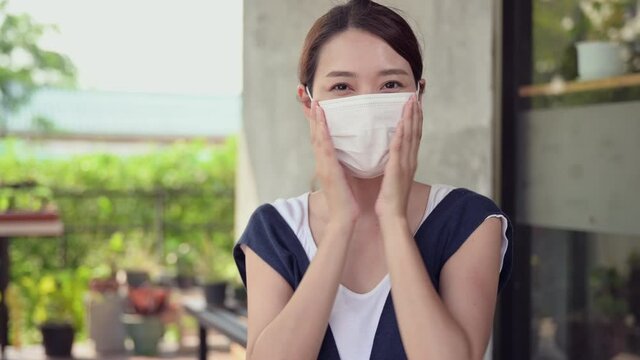 Happy Asian woman sitting smile and putting white medical mask on face for protective infection of coronavirus disease or covid-19 and showing thumb up at home. New normal lifestyle during quarantine.