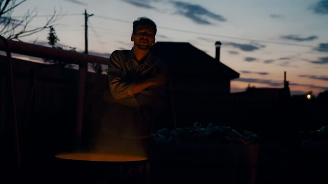A European man in jean jacket near a burning garbage can on his face.