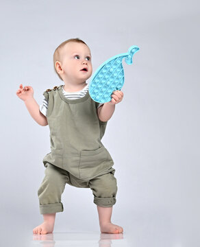 Toddler todler stands on a light background of the studio in a green sandpit overalls and holds a popular toy pop it antistress in his hands.