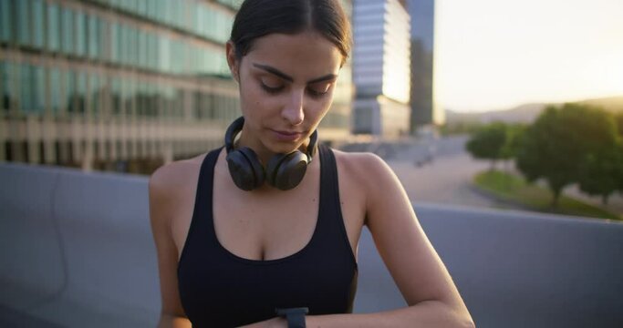Fitness woman takes break after physical activity checks pulse or heart rate on modern smartwatch has running exercise in urban place intense cardio training rapid breathing uses mobile gadgets