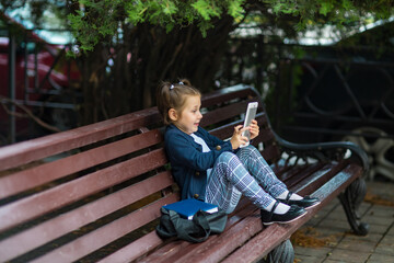 a little girl in a school uniform, sitting on a bench, and holding a tablet in her hands