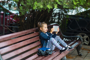 a little girl in a school uniform, sitting on a bench, and holding a tablet in her hands