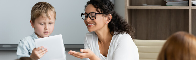 cheerful african american teacher pointing at digital tablet near schoolboy, banner