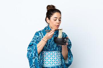 Woman wearing kimono over isolated white background holding a bowl of noodles with chopstick sand blowing it because they are hot