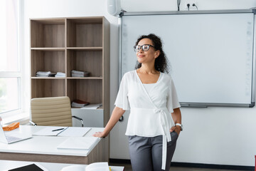 smiling african american teacher standing with hand in pocket in classroom