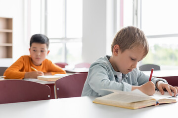 schoolboy writing during lesson near asian classmate on blurred background
