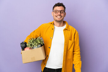 Young caucasian man making a move while picking up a box full of things isolated on purple...