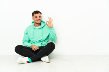 Young handsome caucasian man sitting on the floor showing ok sign with fingers