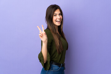 Teenager Brazilian girl over isolated purple background smiling and showing victory sign