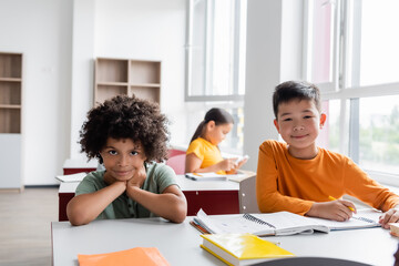 Fototapeta premium multiethnic classmates looking at camera near notebooks in classroom