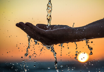 primer plano de una mano cayéndole un chorro de agua con un fondo de atardecer 